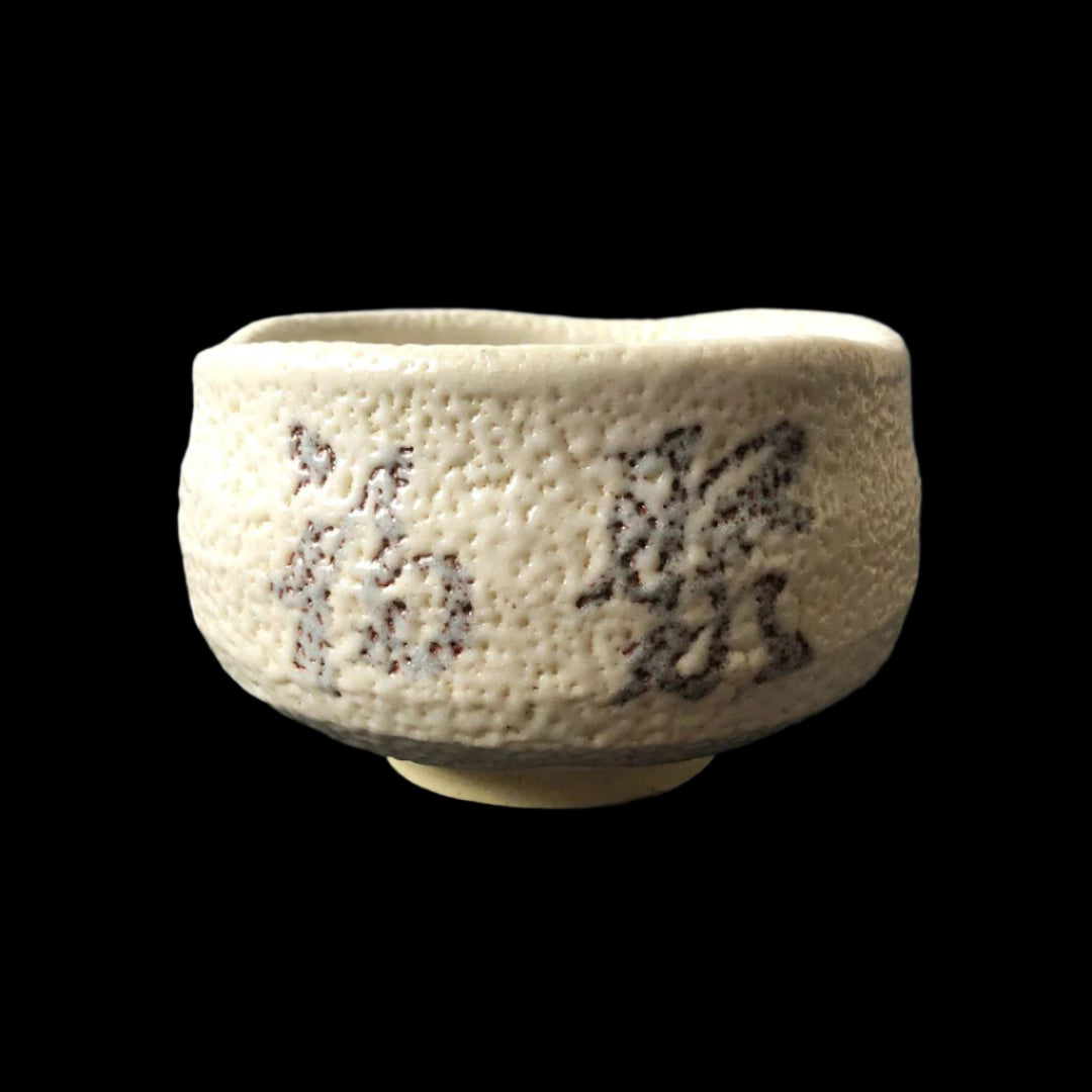 A textured Shino ware Japanese tea bowl with a white feldspar glaze, featuring the kanji for 'Fukuju' meaning 'Many merits that bring happiness' in a subtle grey tone. The tea bowl has an irregular rim and a creamy white finish, with the calligraphy adding an artistic touch. It is set against a dark background, emphasizing its simple yet elegant design.