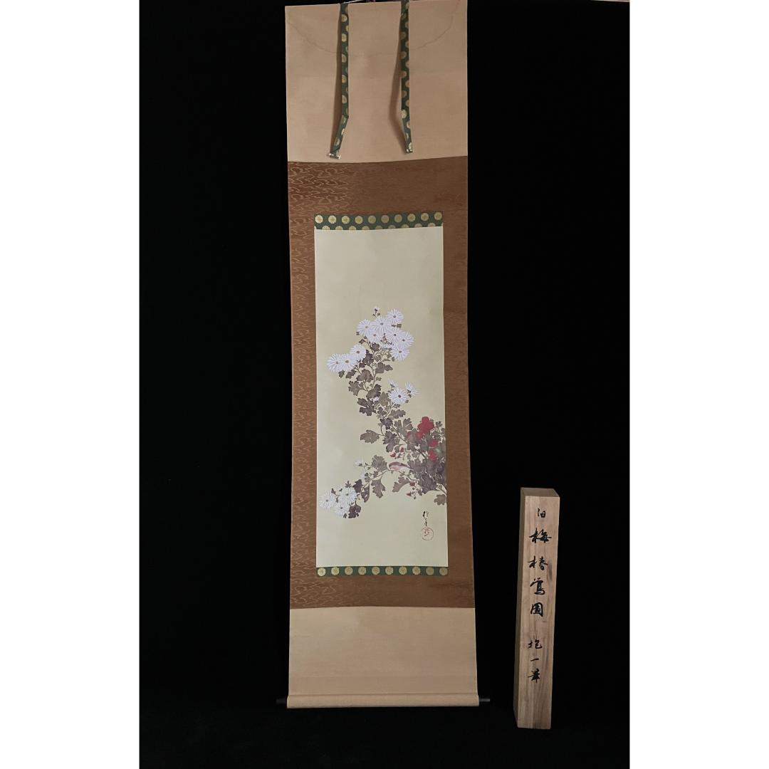 A hanging scroll with a floral painting, bordered by patterned fabric, displayed next to a wooden plank with Japanese characters, all against a black background.