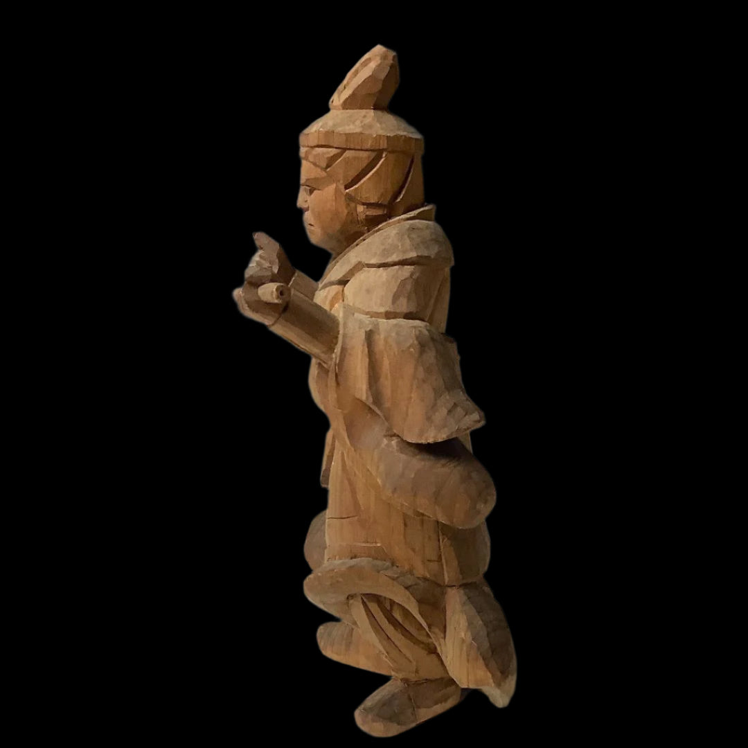 A close-up of a Japanese wood carving depicting Komokuten, a Buddhist guardian deity, with a solemn expression, clad in layered robes, and headdress, gesturing with both hands.
