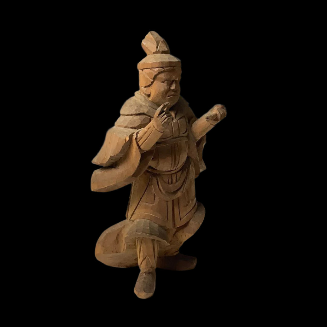 A close-up of a Japanese wood carving depicting Komokuten, a Buddhist guardian deity, with a solemn expression, clad in layered robes, and headdress, gesturing with both hands.