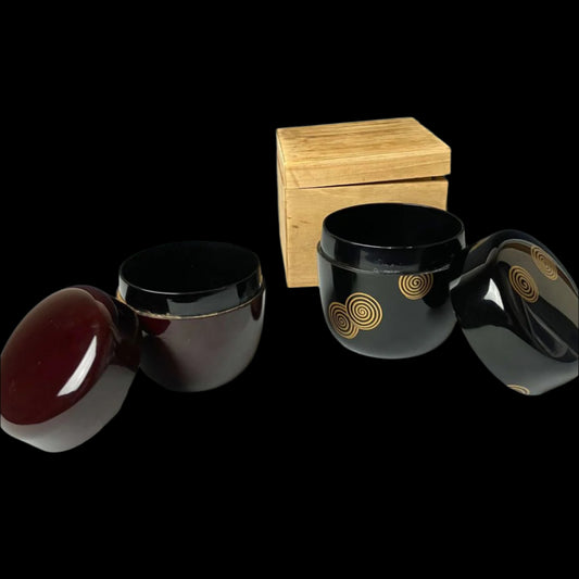 Two open 19th-century Japanese lacquered makie natsume tea caddies, one in deep brown and the other in black with gold spiral designs, accompanied by a wooden box, highlighting their traditional craftsmanship and elegant design.