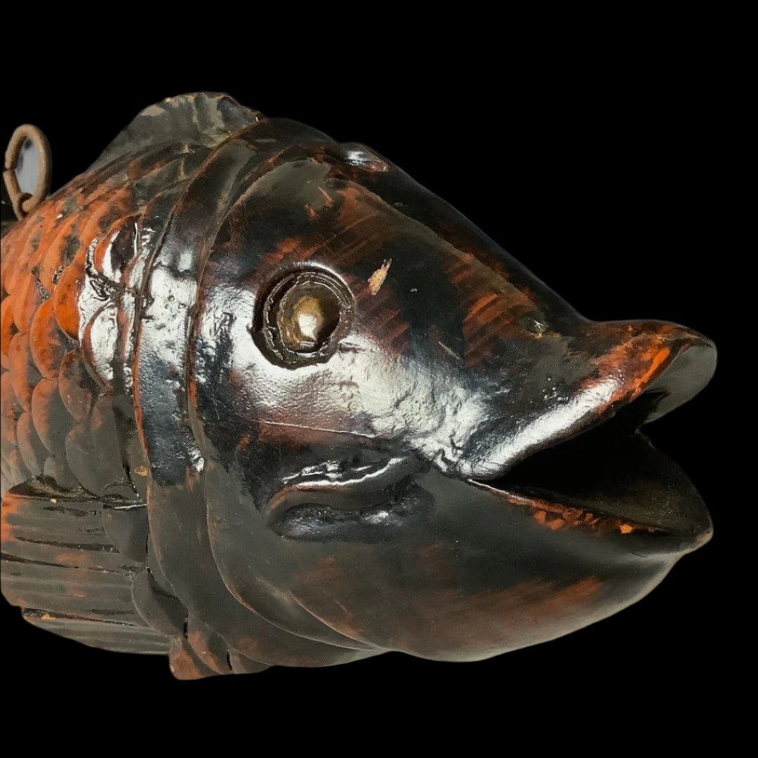 A close-up of a sculpted wooden fish head with a dark, glossy finish, featuring detailed eyes, mouth, and scale texture, with a metal ring attached above, set against a gradient gray backdrop.