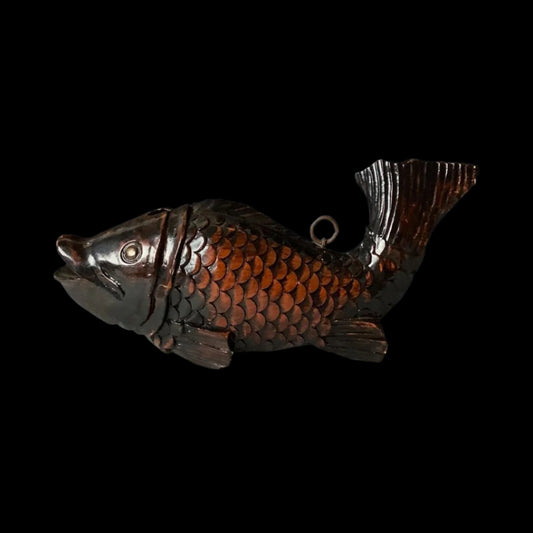A decorative, dark brown, wooden fish sculpture with detailed scales and fins, featuring a metallic ring on its back for hanging, displayed on a gray surface against a gradient gray background.