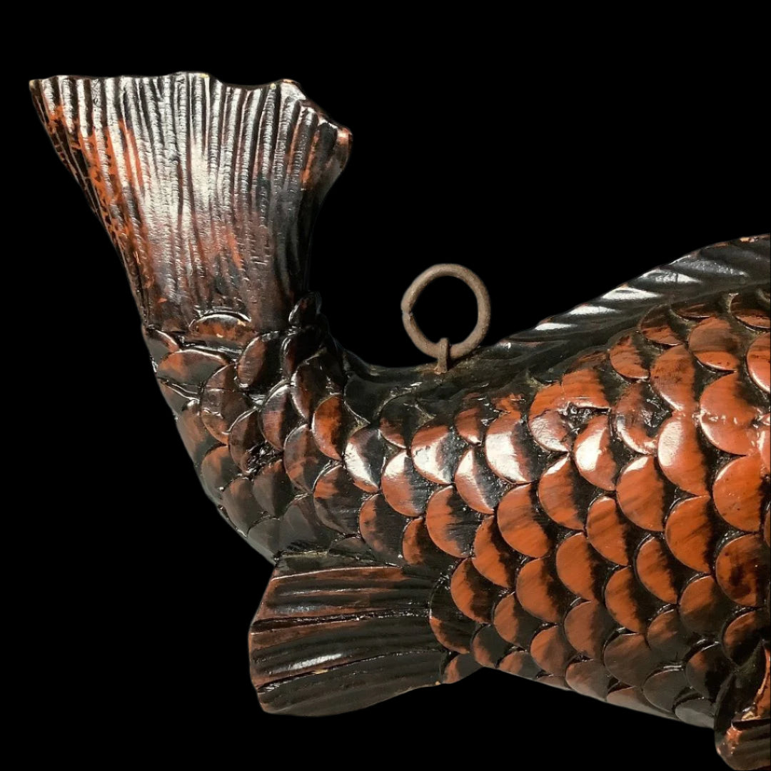 A side view of a wooden fish sculpture with a striking tail fin and detailed scales, finished in dark glossy brown with highlights. A metal hanging ring is attached near the tail fin, against a smooth gradient gray background.