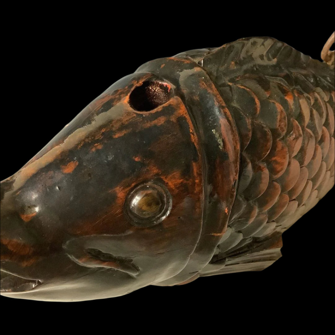 Close-up of the head of a wooden fish sculpture with a shiny, dark patina finish. The fish has a carved eye and mouth, textured scales, and a hole on top where a piece is missing, set against a light gray background with slight scuff marks.