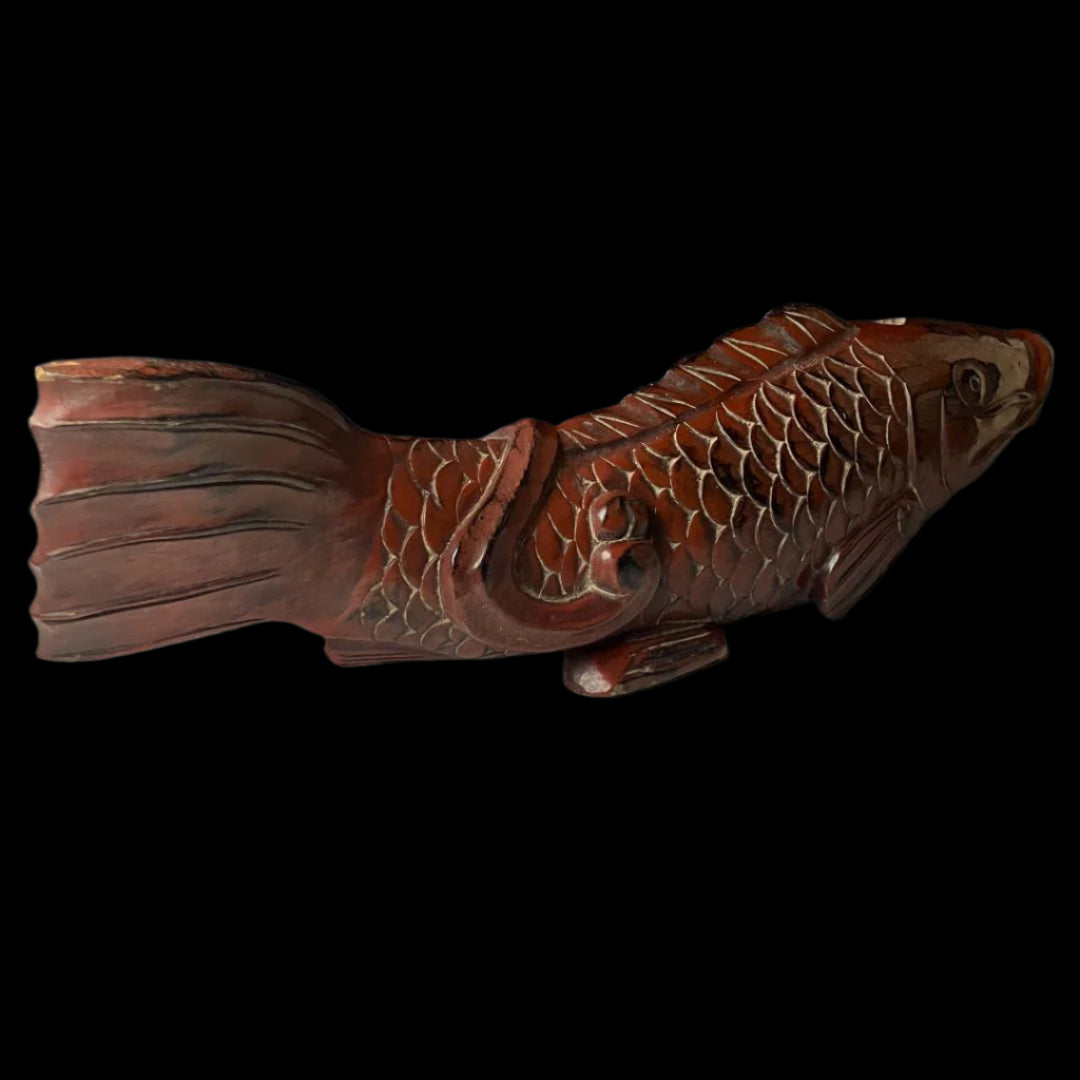 This image displays a full side view of an antique wooden fish carving, with a rich reddish-brown patina, detailed scales, and a gracefully carved tail. The fish's eye is detailed and expressive, adding life to the carving. The object stands on a flat surface against a neutral background, highlighting its artistic qualities and the fine craftsmanship.