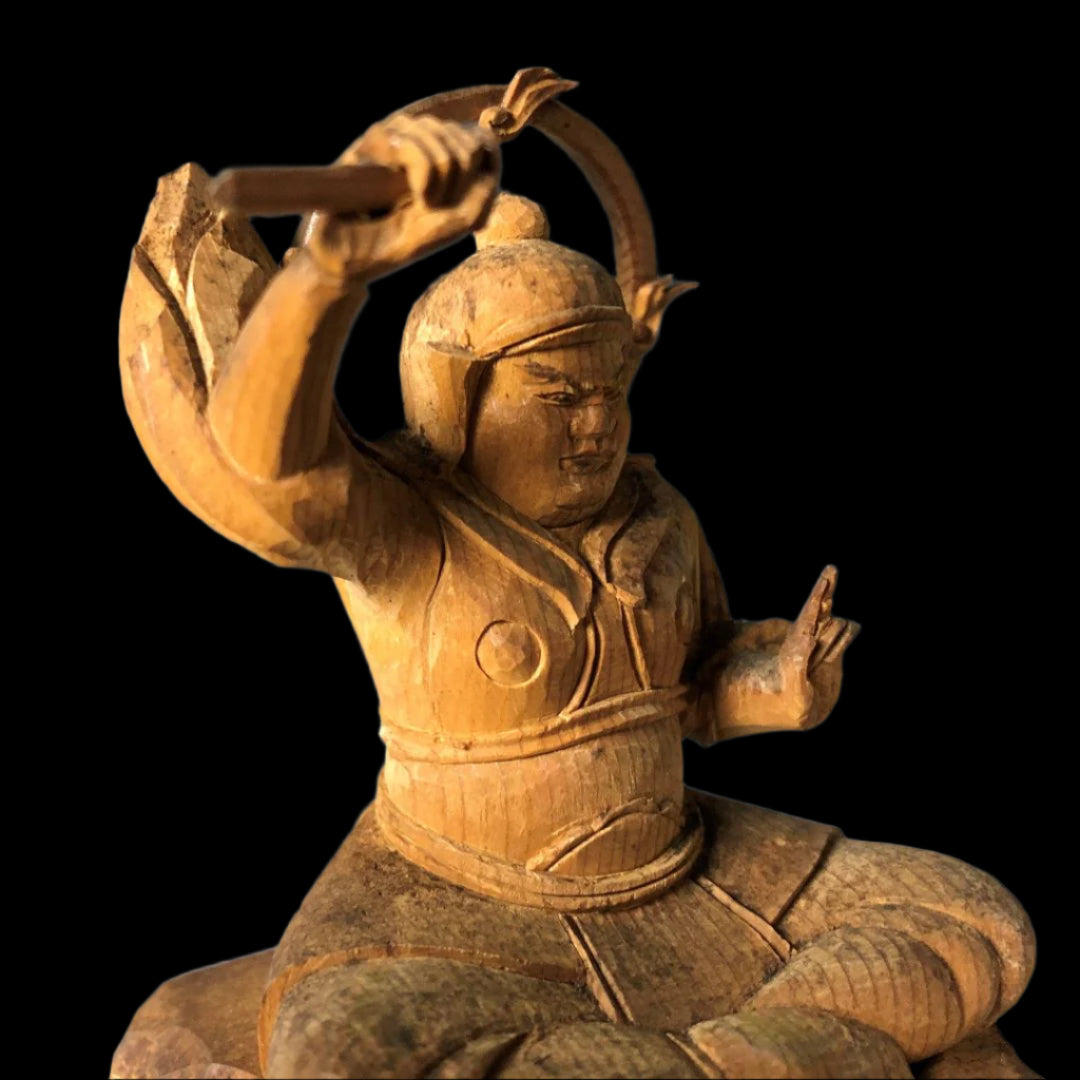 A wooden sculpture of a seated figure with a helmet, pointing upwards, holding a curved object, showcasing detailed traditional attire and expressive craftsmanship.