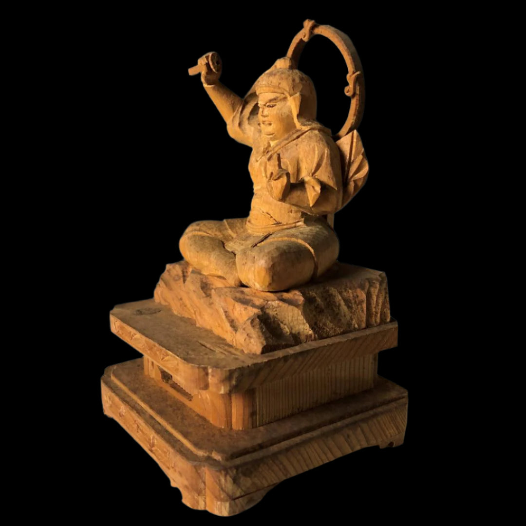 A wooden sculpture of a joyful figure seated cross-legged, with one arm raised and holding a ring, on a multi-tiered wooden base. The figure is carved with visible smiles and intricate detailing on the clothing and base.