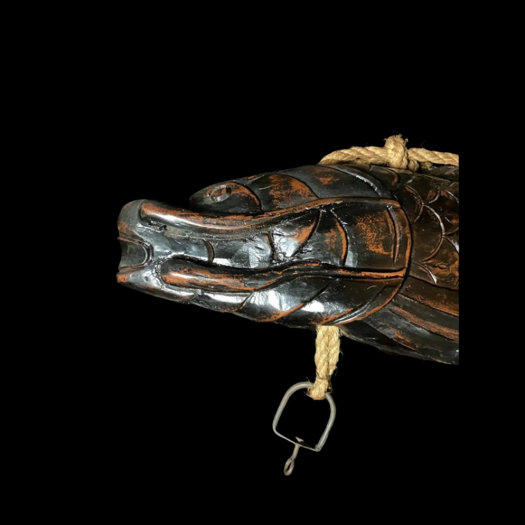A carved wooden fish head is being used as a pot carrier. The fish head has intricate carvings and a hook, with a thick rope passing through its head. It is placed on a black cloth, showcasing its unique features.