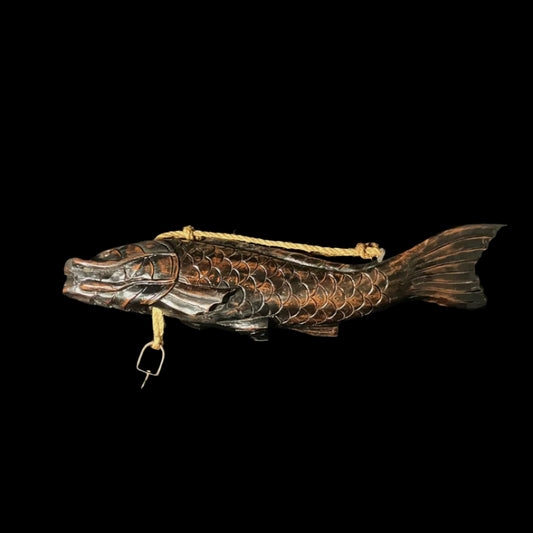 A wooden fish with intricate carvings is being used as a pot carrier. The fish has a hook and a thick rope passing through its body. It is placed on a black cloth, highlighting its unique features.