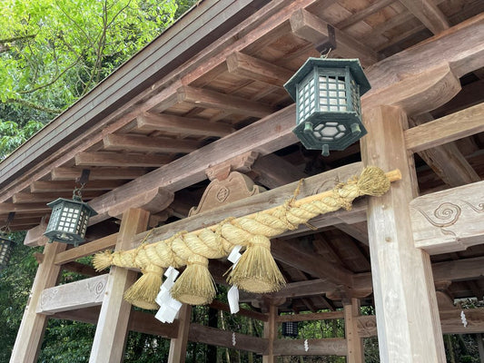 A section of a Japanese shrine with wooden architecture, hanging metal lanterns, and a sacred shimenawa rope with white paper strips, set against a backdrop of green leaves.