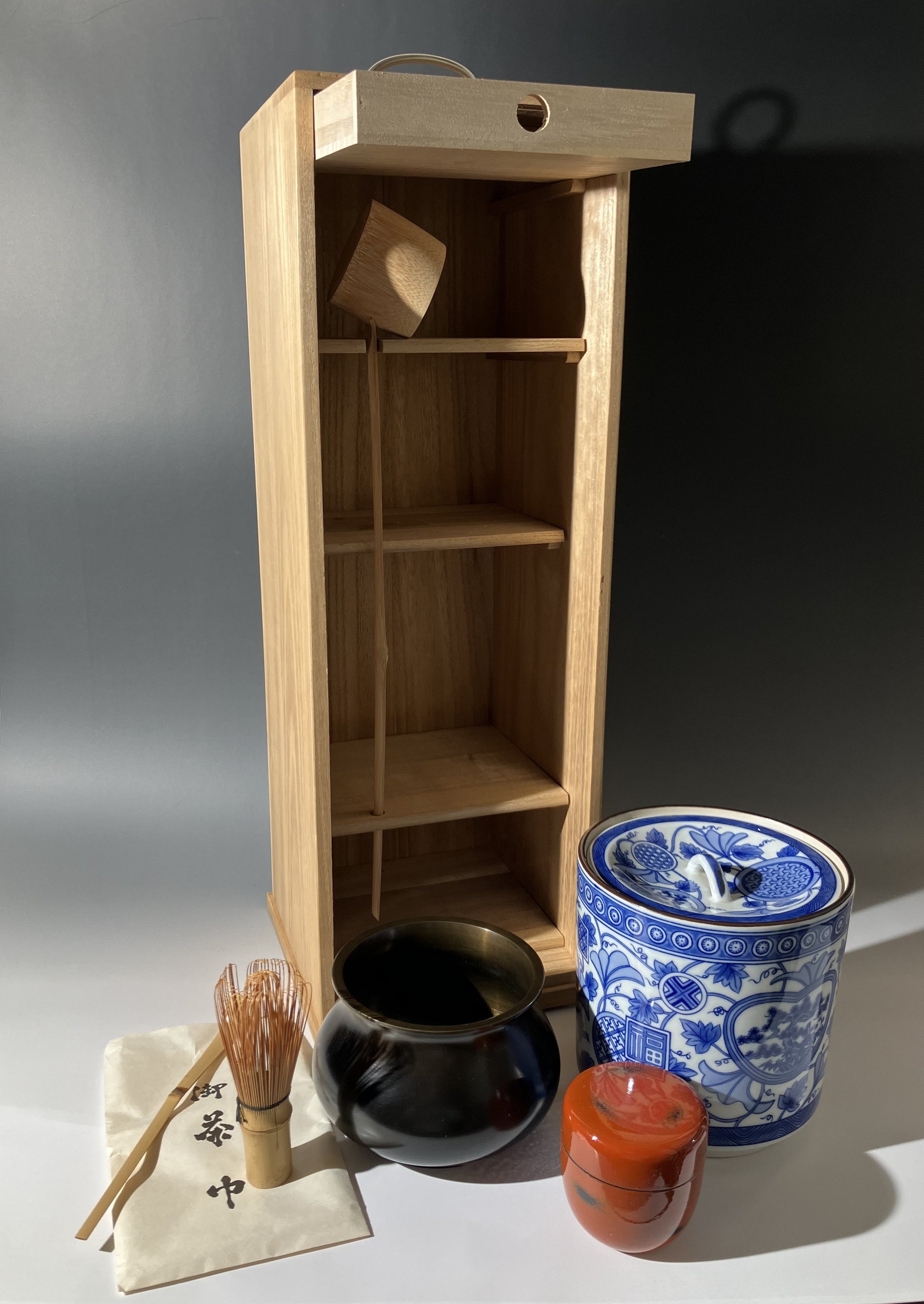 Wooden shelf with tea-making items including a blue and white teapot, black cup, and red candle on a gray background.