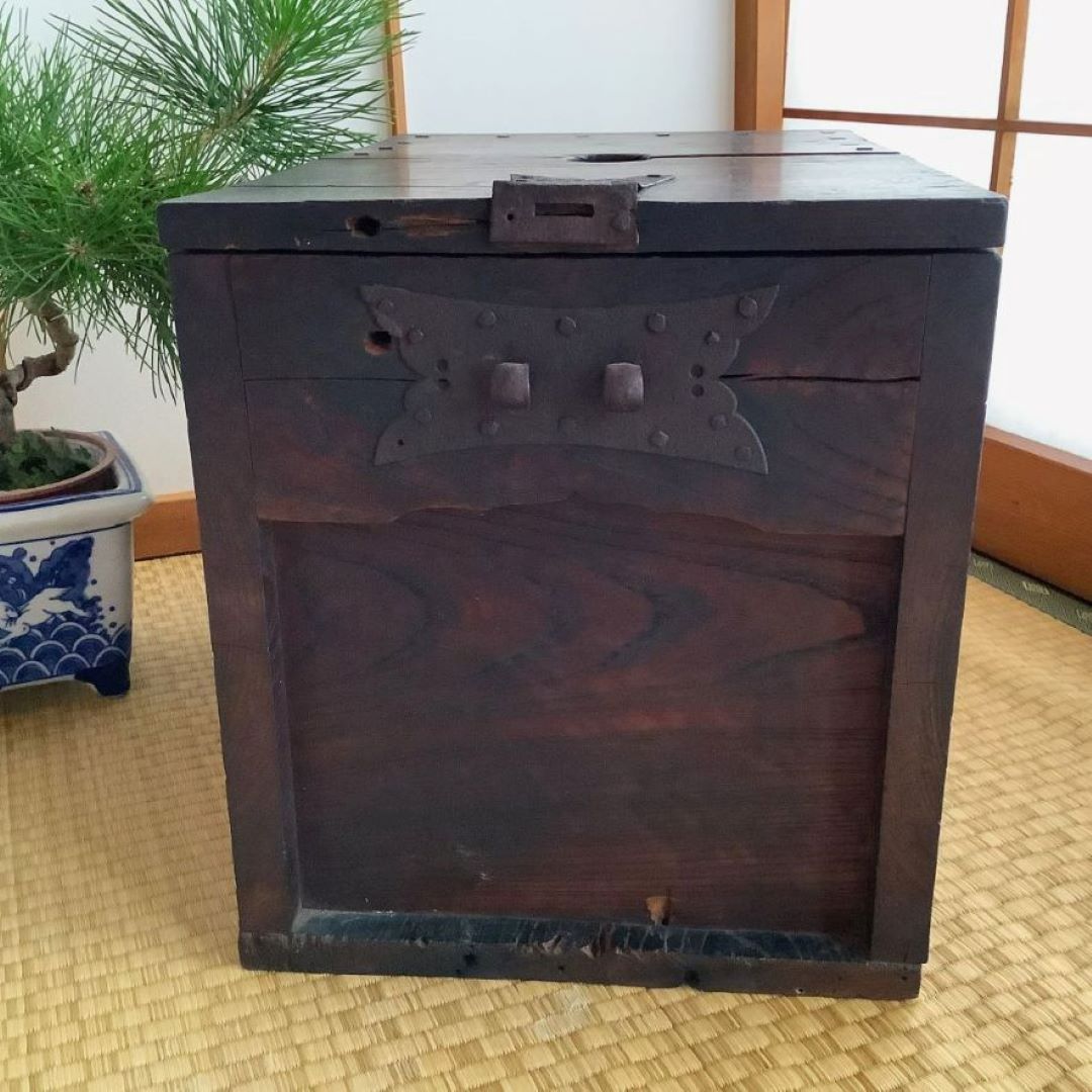 A close-up view of a vintage Japanese wooden chest on a tatami mat floor. The chest features a dark stain and is adorned with traditional metal hardware, including a large decorative hinge and square metal accents. To the left, a portion of a bonsai tree can be seen, resting in a blue and white ceramic pot, adding a touch of nature to the composition. The room is lit with natural light, suggesting a peaceful interior setting.