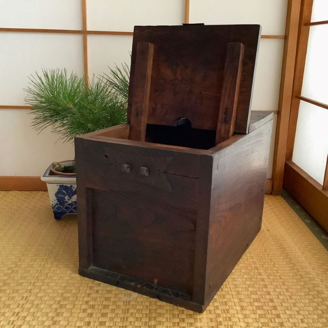 An open antique Japanese wooden chest is placed on a tatami mat. The lid of the chest is propped open, revealing the dark interior. On the left, a bonsai tree in a blue and white pot is partially visible, and in the background, the room is divided by a shoji screen, which allows natural light to filter through and create a warm, inviting atmosphere. The chest's aged wood and traditional metal hardware emphasize its rustic charm and cultural heritage.