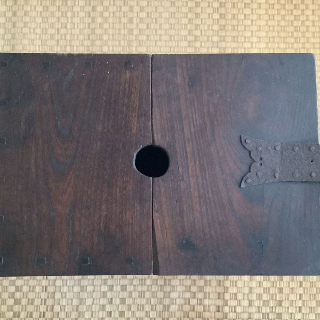 The top view of an antique Japanese wooden chest's lid, laid flat open and showing a central circular hole for a handle. The dark wood of the lid contrasts with the light tatami mat beneath it. The visible side of the lid displays traditional metal hardware with an intricate design, indicative of its cultural craftsmanship.