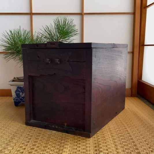 A traditional Japanese wooden chest with metal hardware is sitting on a tatami floor mat. Behind the chest are two small pine branches in a decorative arrangement, with a shoji screen as a backdrop creating a serene and culturally rich atmosphere. The chest appears antique, with a rich patina on the wood and ornate metal accents, suggesting it may be a piece of heritage furniture.