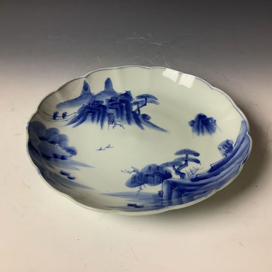  A traditional Japanese blue and white porcelain plate with a scalloped edge, depicting a serene mountainous landscape with trees and small figures, reflecting the classic Asian blue-ink art style.