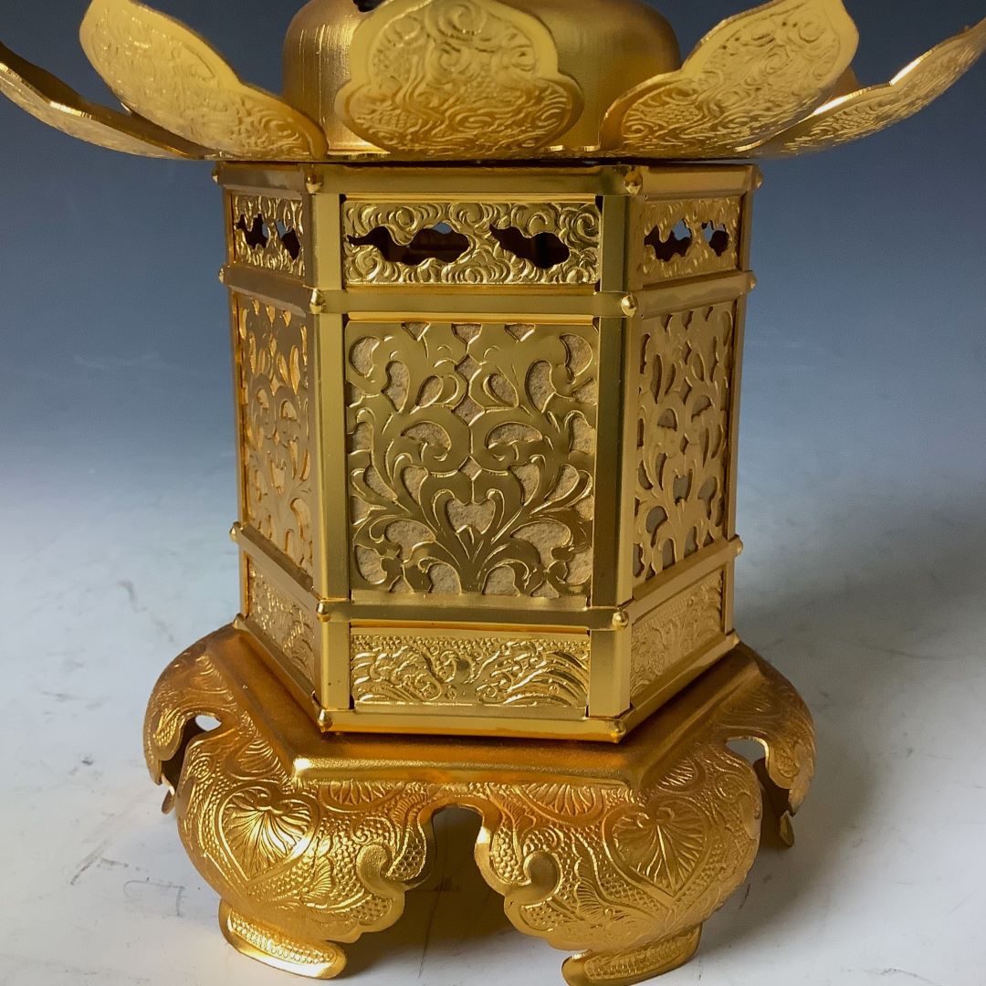 Close-up of a golden Japanese lantern with elaborate floral patterns and wing-like embellishments, resting on a carved base against a dark background.