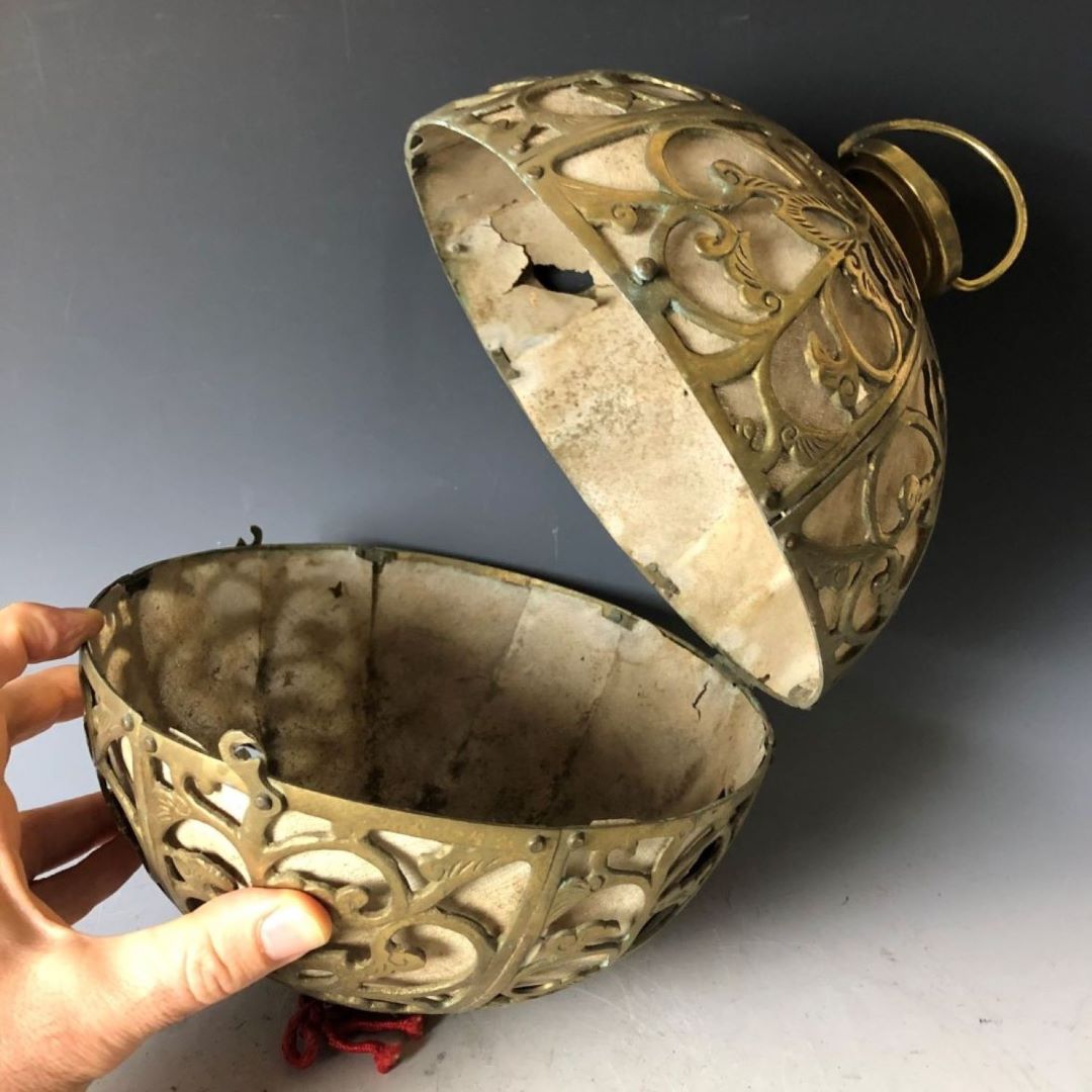 A hand is opening a brass spherical lantern with ornate patterns to reveal its hollow interior, against a gradient gray background.