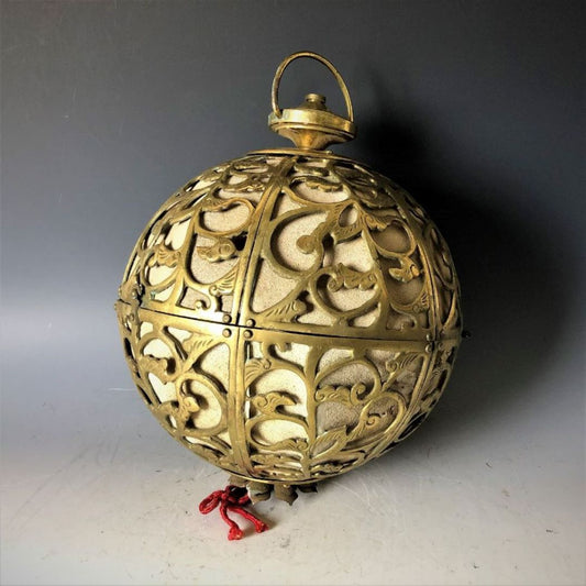 A decorative brass orb with intricate cut-out patterns, featuring a loop on top for hanging and standing on a small base with a red tassel, against a gradient gray background.