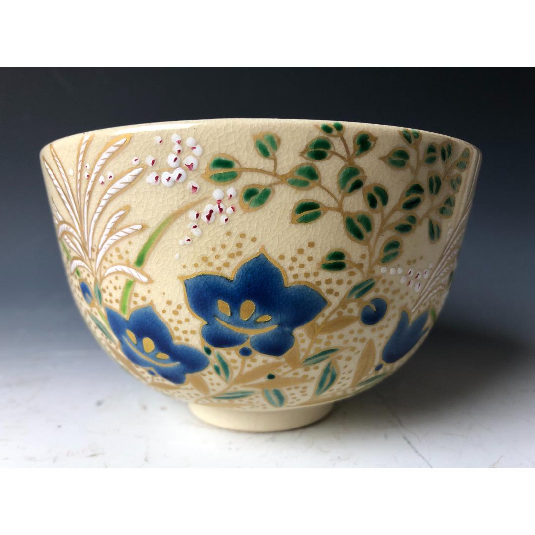 A Japanese Chawan tea bowl with a cream-colored crackled glaze, showcasing a vibrant hand-painted design of blue flowers, green leaves, and delicate pink dots that appear to be cherry blossoms, indicative of traditional Japanese aesthetics.