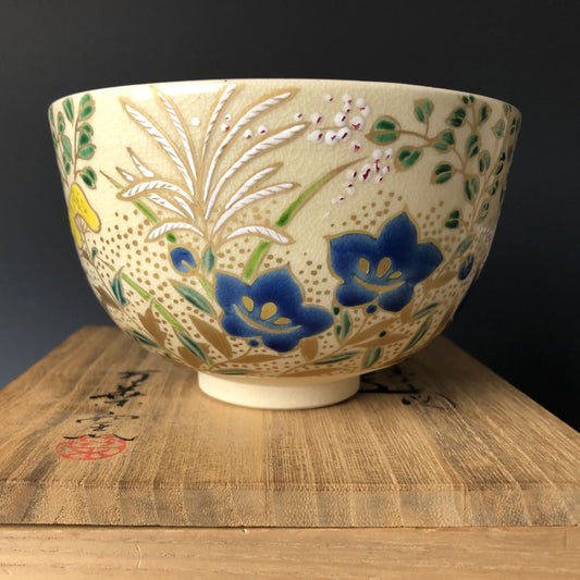 A Kiyomizu-yaki Chawan tea bowl with intricate floral patterns in yellow and blue, resting on a wooden stand with Japanese calligraphy, exemplifying traditional Kyoto ceramics craftsmanship.