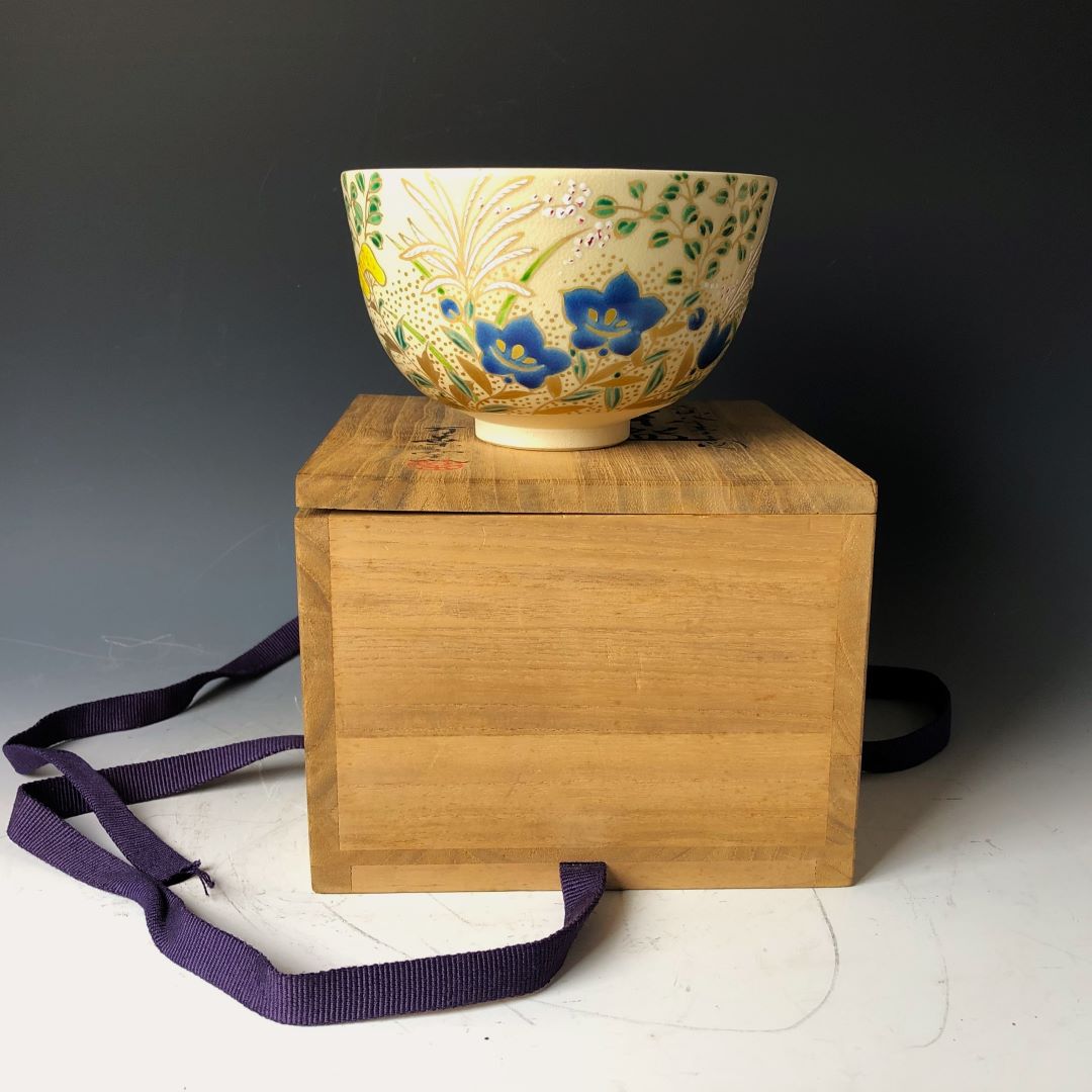 Image of a Japanese Chawan tea bowl with blue floral patterns on a light cream background, displayed atop a wooden box with a dark purple fabric strap, against a dark backdrop, highlighting its traditional beauty and craftsmanship.