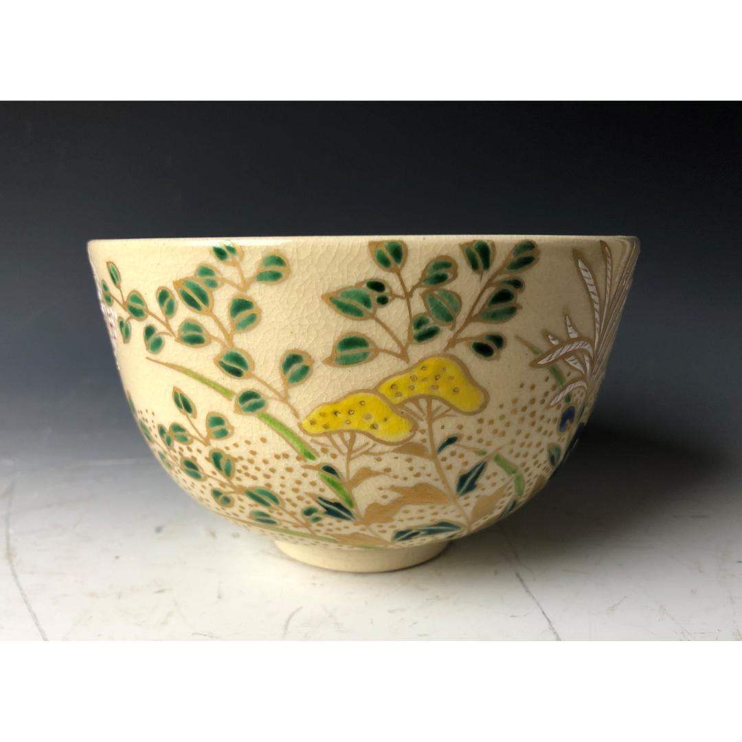 This image shows a cream-colored Japanese Chawan tea bowl, adorned with hand-painted yellow and green botanical motifs, showcasing the traditional Japanese artistry in ceramic design, against a muted background.