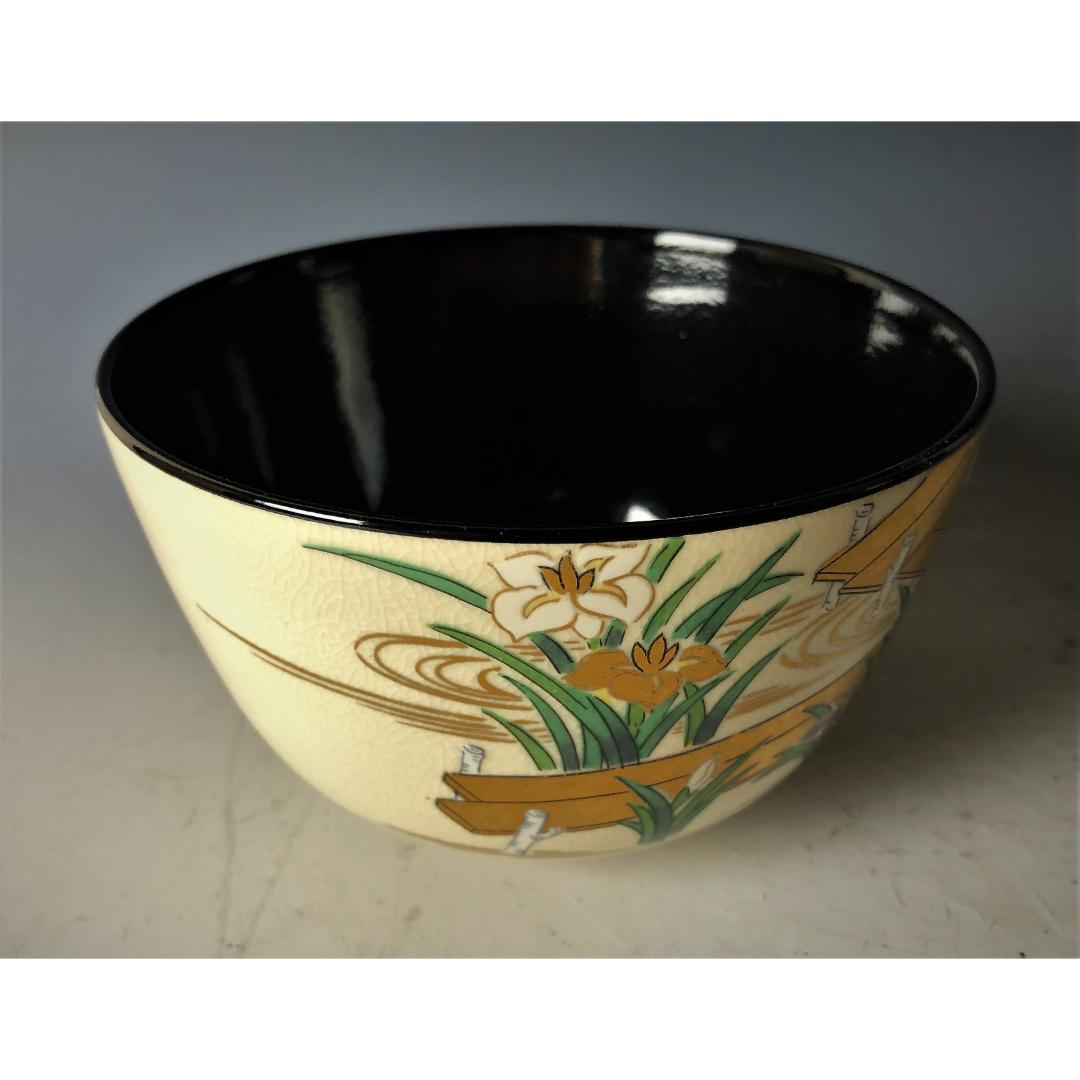 Angled view of a Kyo-yaki tea bowl with a shiny black interior and a hand-painted exterior featuring golden and orange irises against an ivory crackled background.