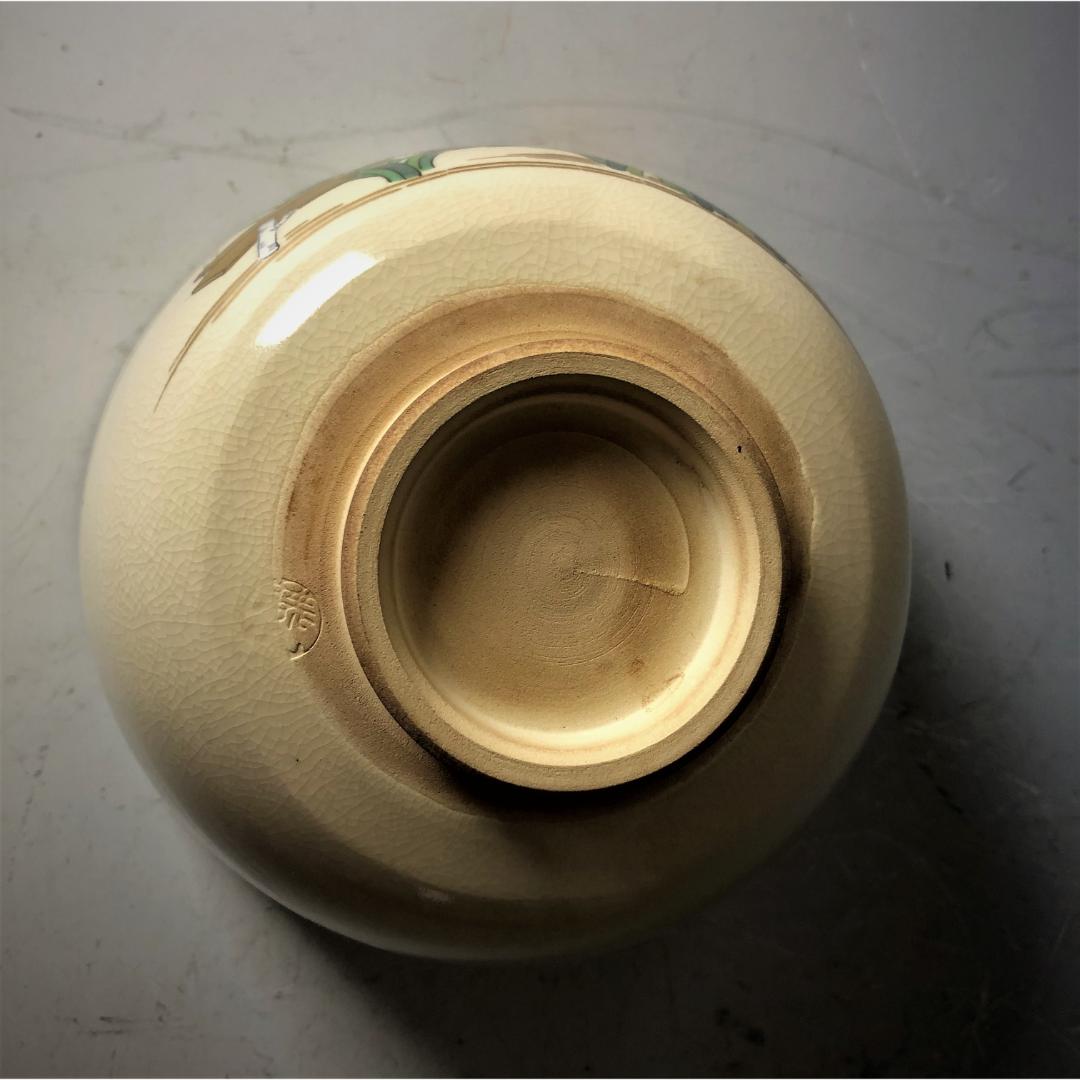  Overhead view of the bottom of a Kyo-yaki tea bowl, displaying the textured, crackled ivory glaze and the maker's mark, set on a grey surface with a hint of the bowl's painted exterior.