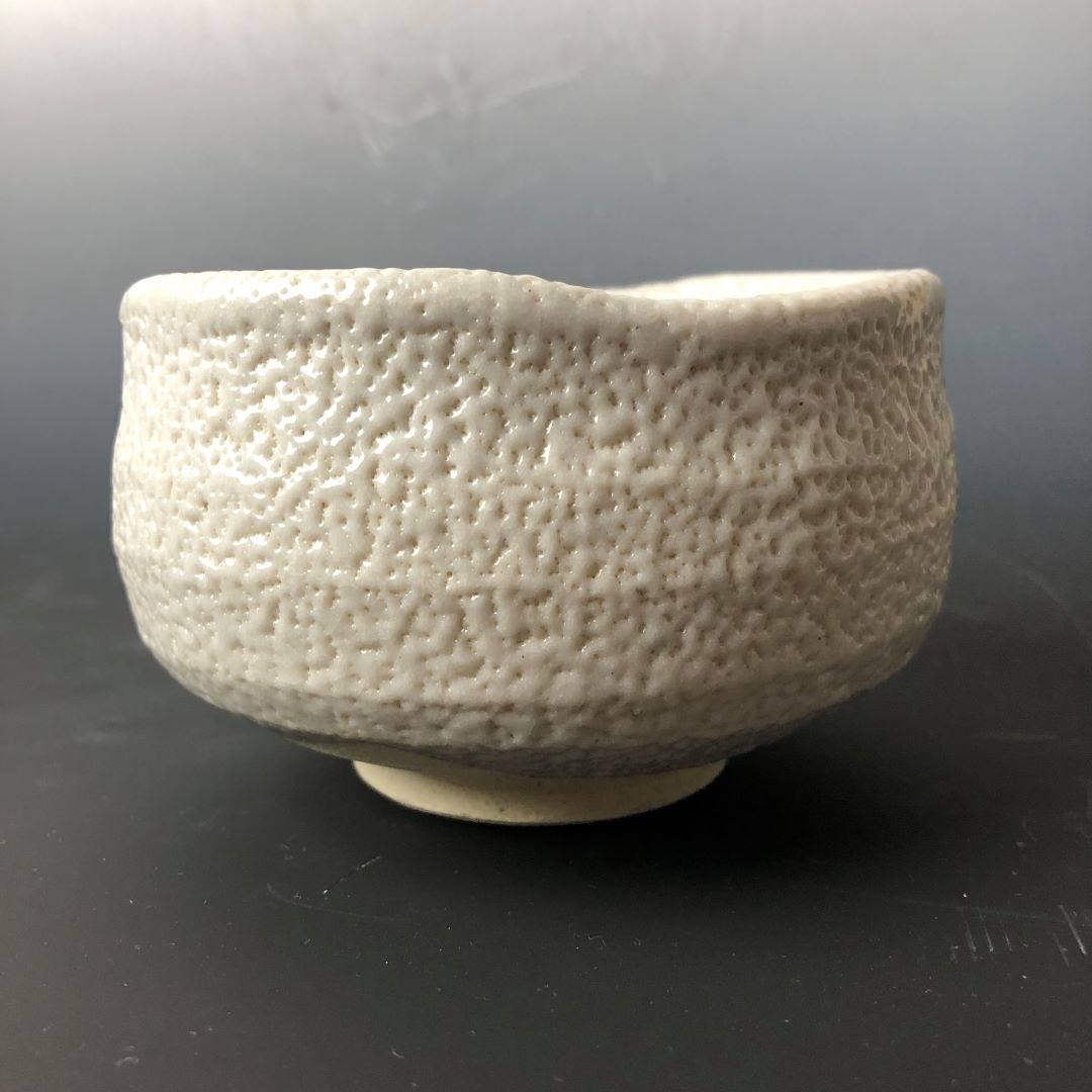 Side view of a textured white Shino ware Japanese tea bowl on a dark background.