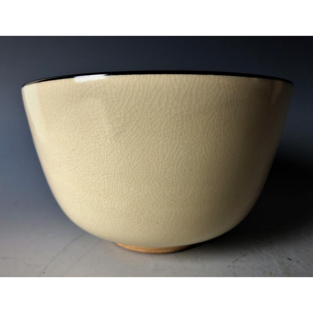 A simple yet elegant side view of a Kyo-yaki tea bowl, displaying a crackled ivory glaze with a contrasting black rim, on a gradient grey background.