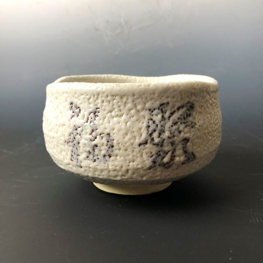 A textured Shino ware Japanese tea bowl with a white feldspar glaze, featuring the kanji for 'Fukuju' meaning 'Many merits that bring happiness' in a subtle grey tone. The tea bowl has an irregular rim and a creamy white finish, with the calligraphy adding an artistic touch. It is set against a dark background, emphasizing its simple yet elegant design.