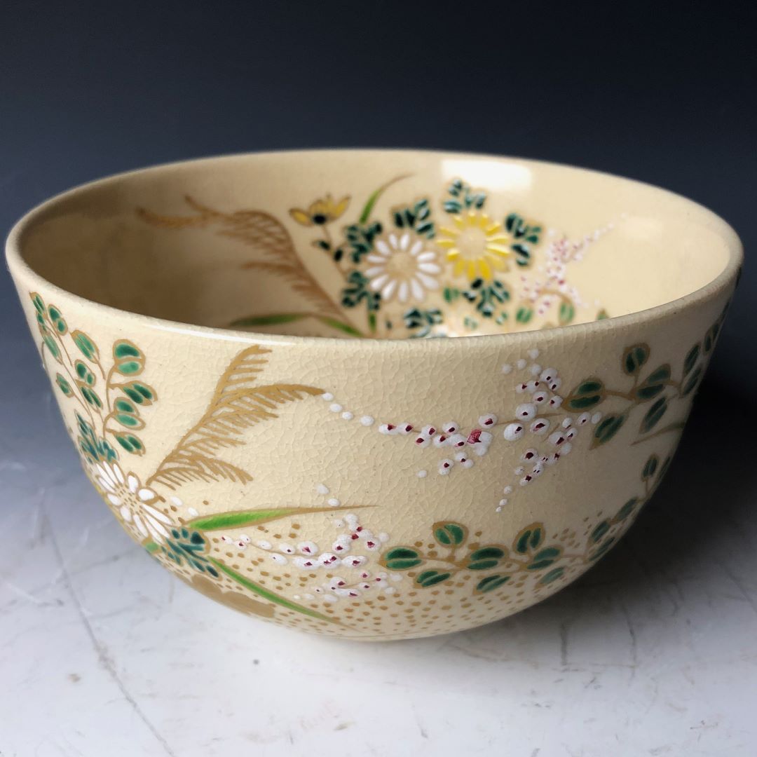 A hand-painted Japanese Chawan tea bowl, adorned with a cream-colored glaze and intricate floral designs featuring yellow daisies, green foliage, and delicate white blossoms with red centers. The bowl's textured surface adds depth to the vibrant, detailed artwork.