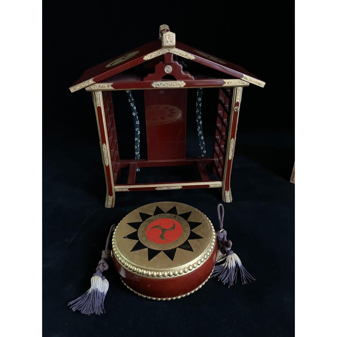 Frontal view of a traditional Japanese Shinto drum with a bold blue and white spiral design on the drumhead, edged with gold studs on the burgundy base, set against the backdrop of its wooden temple stand.