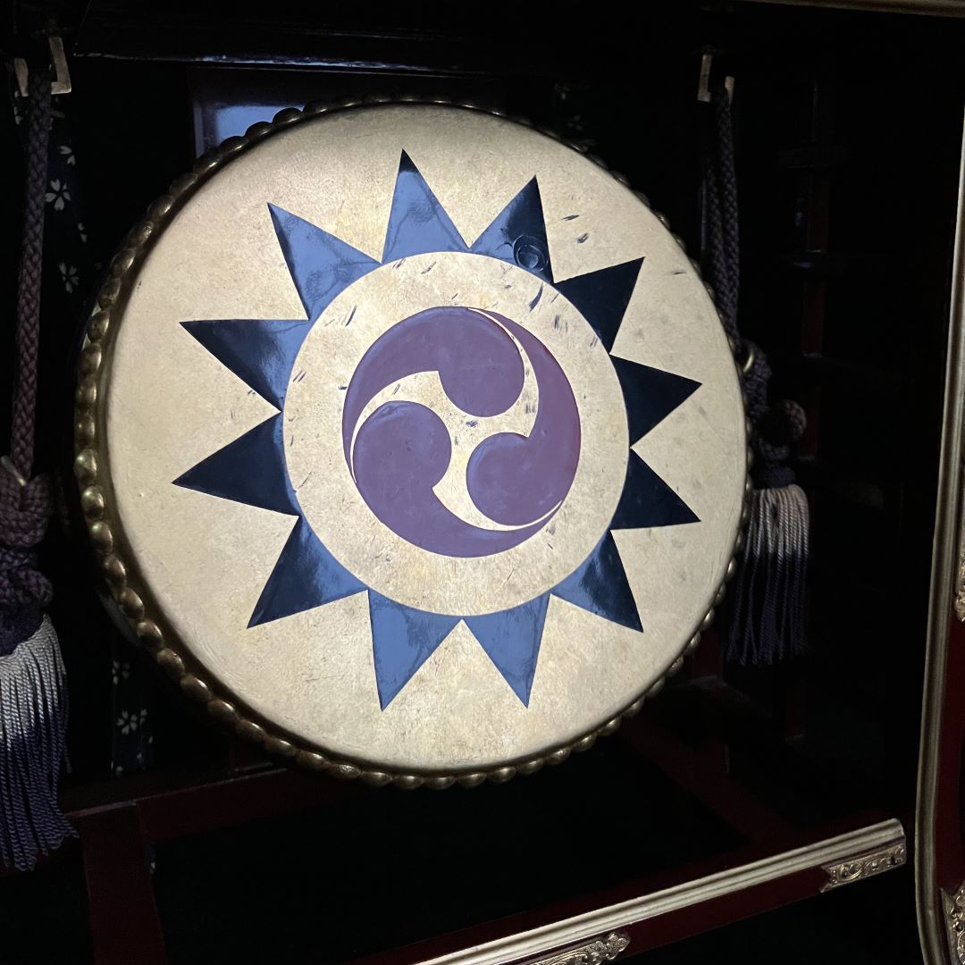 Top-down view of a Japanese Shinto drumhead, featuring a central purple and white swirl design within a blue starburst pattern on a textured beige background.