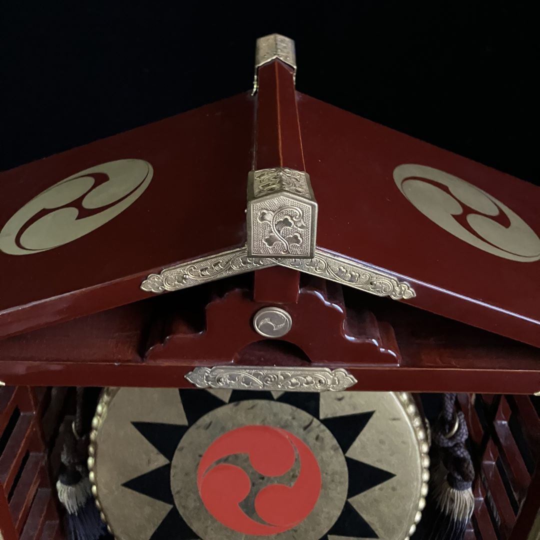 Close-up of the decorative peak of a Japanese Shinto drum’s temple roof, showing engraved floral motifs on the silver metalwork against the burgundy wood.