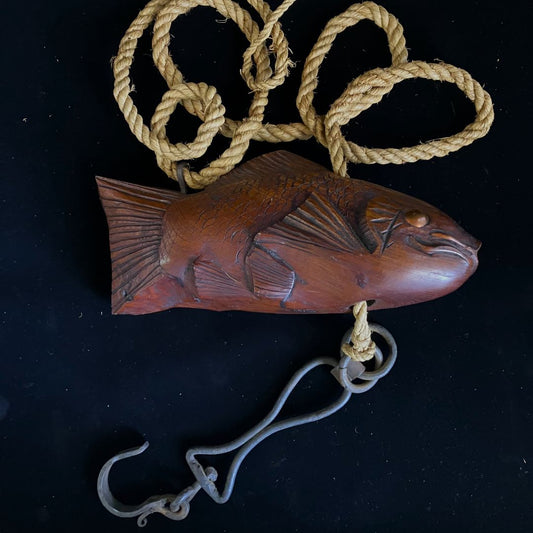 A wooden fish sculpture, possibly used as a jizaikagi or hearth hook, with detailed scales and fins, alongside a metal hook attached to a natural fiber rope with decorative knots. The items are displayed on a black background.