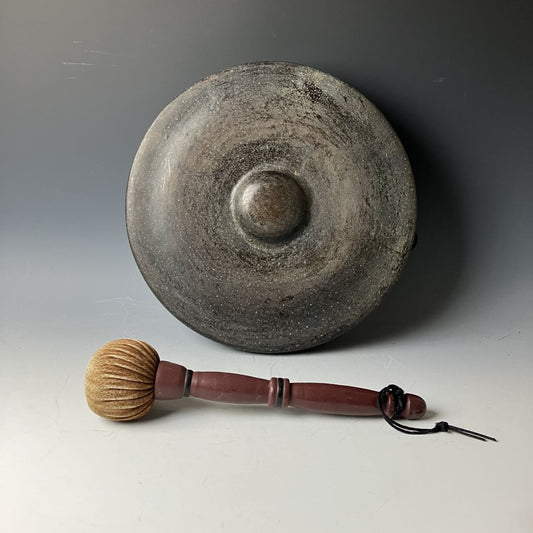 An aged Japanese iron gong with a central bulge and a wooden mallet with a textured head, displayed against a grey background, for meditation or spiritual rituals.