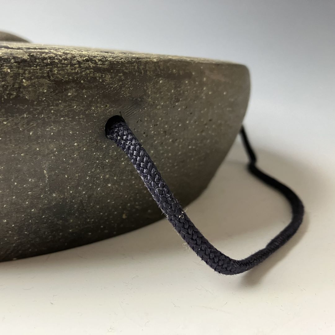 Side view of a weathered Japanese iron gong with a raised center boss, displaying a patinated texture and a dark cord attached, against a light grey backdrop.
