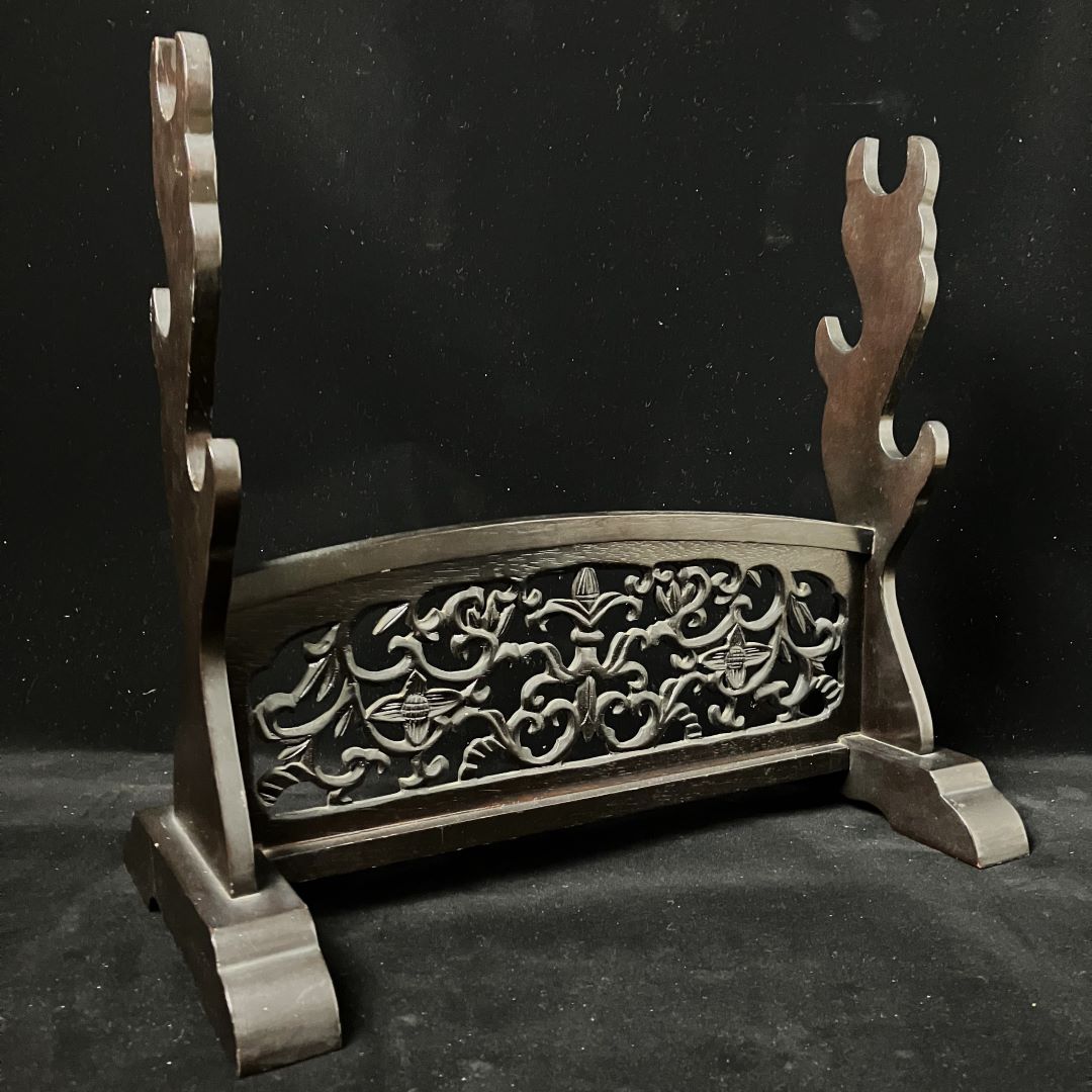 This image displays a wooden katana stand with an openwork design, featuring a curved rest and support arms, on a black backdrop.