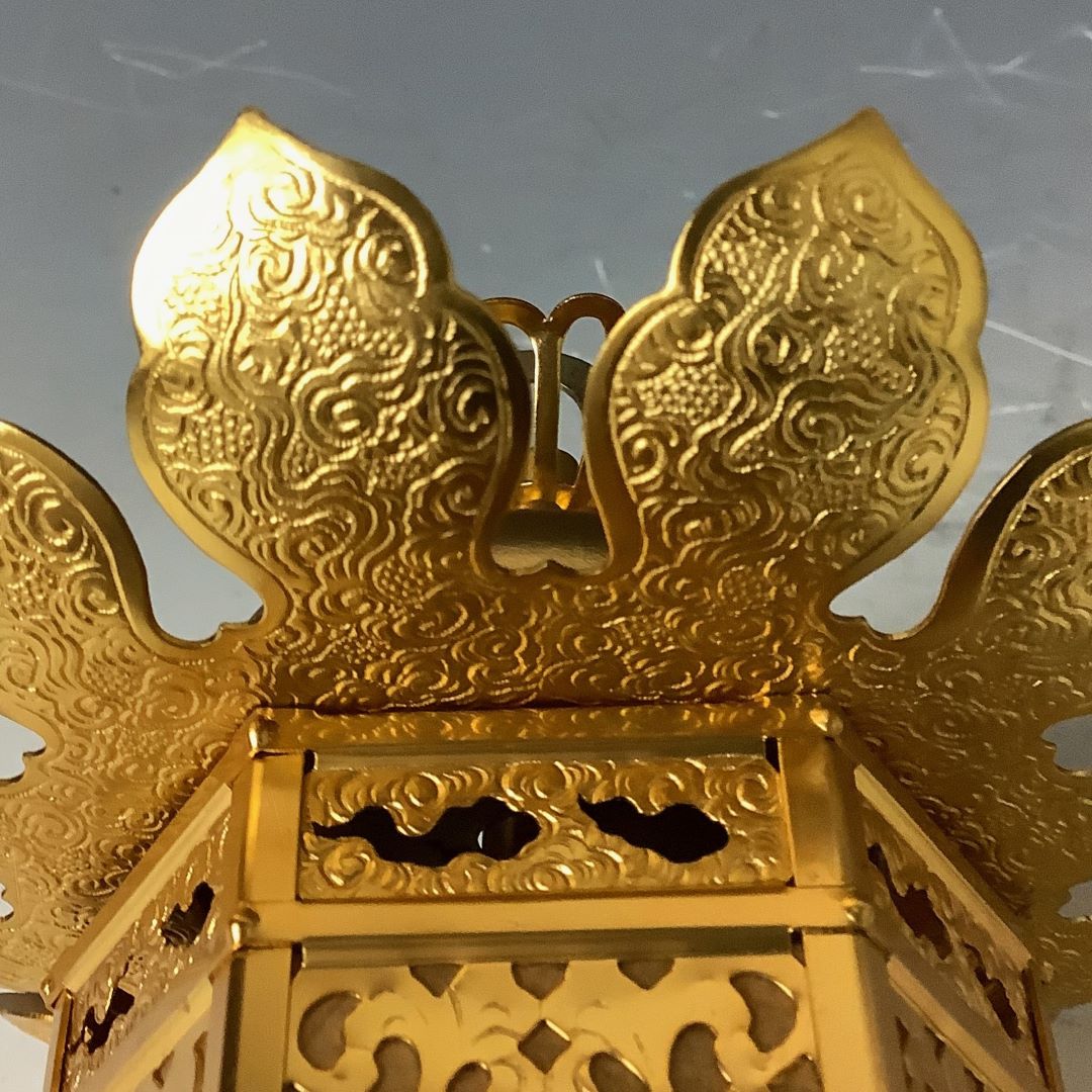 A close-up view of the top of a golden Japanese lantern, featuring ornate, embossed wing-like designs and a decorative handle.