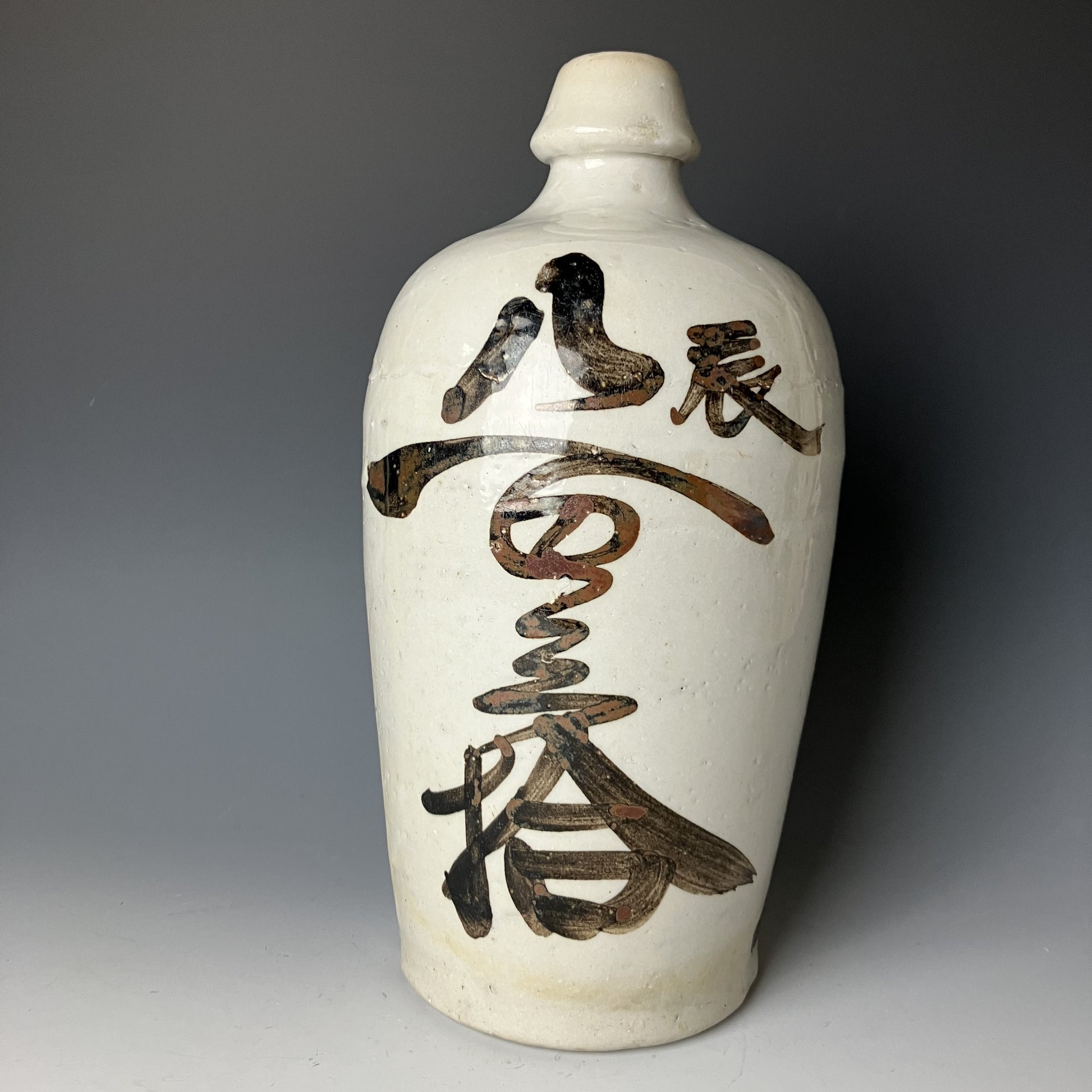A vintage Japanese ceramic sake bottle (Tokkuri) with bold calligraphy, featuring a white background with prominent dark brown and black brush strokes.