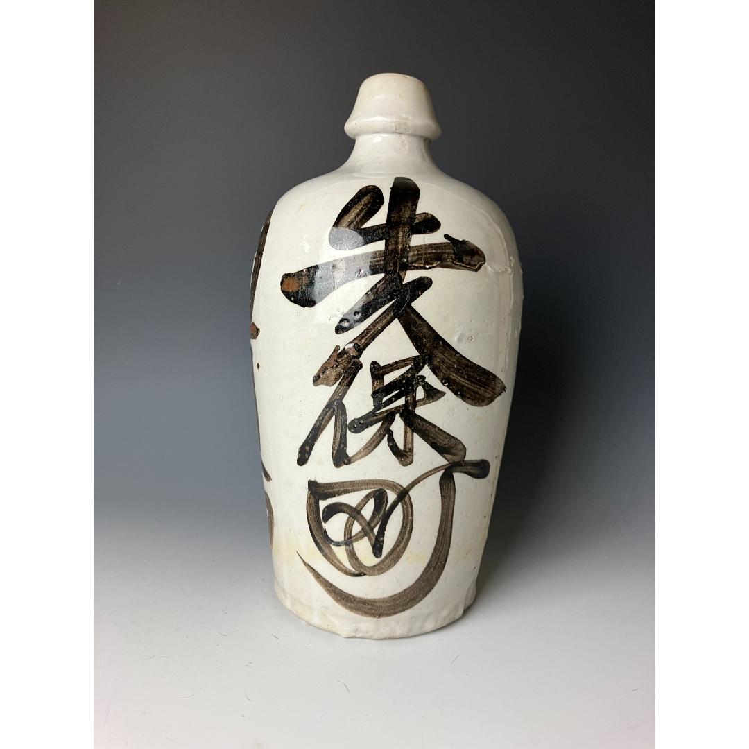 An antique Japanese ceramic sake bottle (Tokkuri) with hand-painted kanji characters in dark brown on a textured white background, displayed from a side angle.