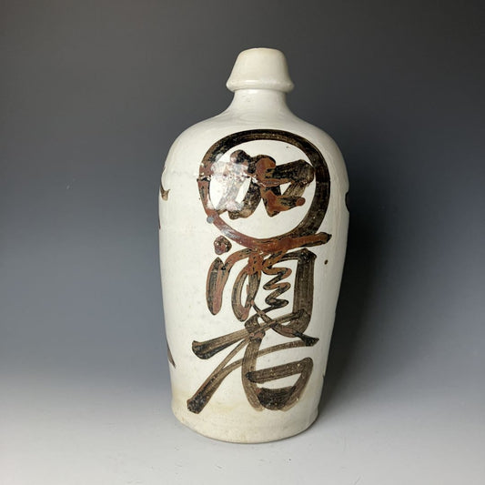 A traditional Japanese ceramic sake bottle (Tokkuri) with calligraphic kanji characters, featuring a white base with brown and black glaze accents, showing signs of age and usage.