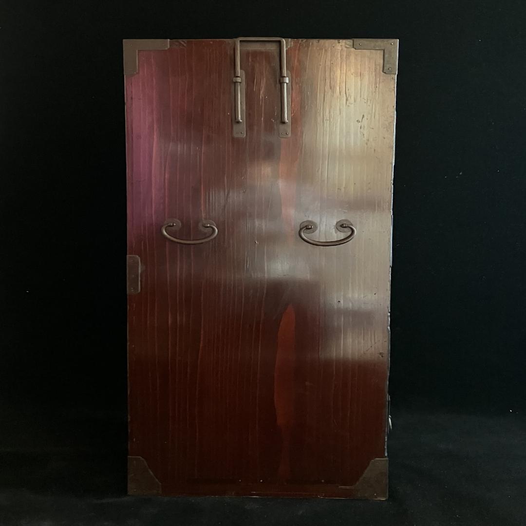 Wooden door of a Japanese tansu cabinet with a warm reddish-brown finish, featuring metal hinges at the top and two curved metal handles, set against a black background.