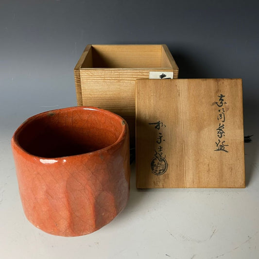 A Japanese Raku yaki tea bowl in Akaraku style, with a rich orange glaze and crackled texture, displayed next to its custom wooden box, which has Japanese calligraphy and a seal on the lid.