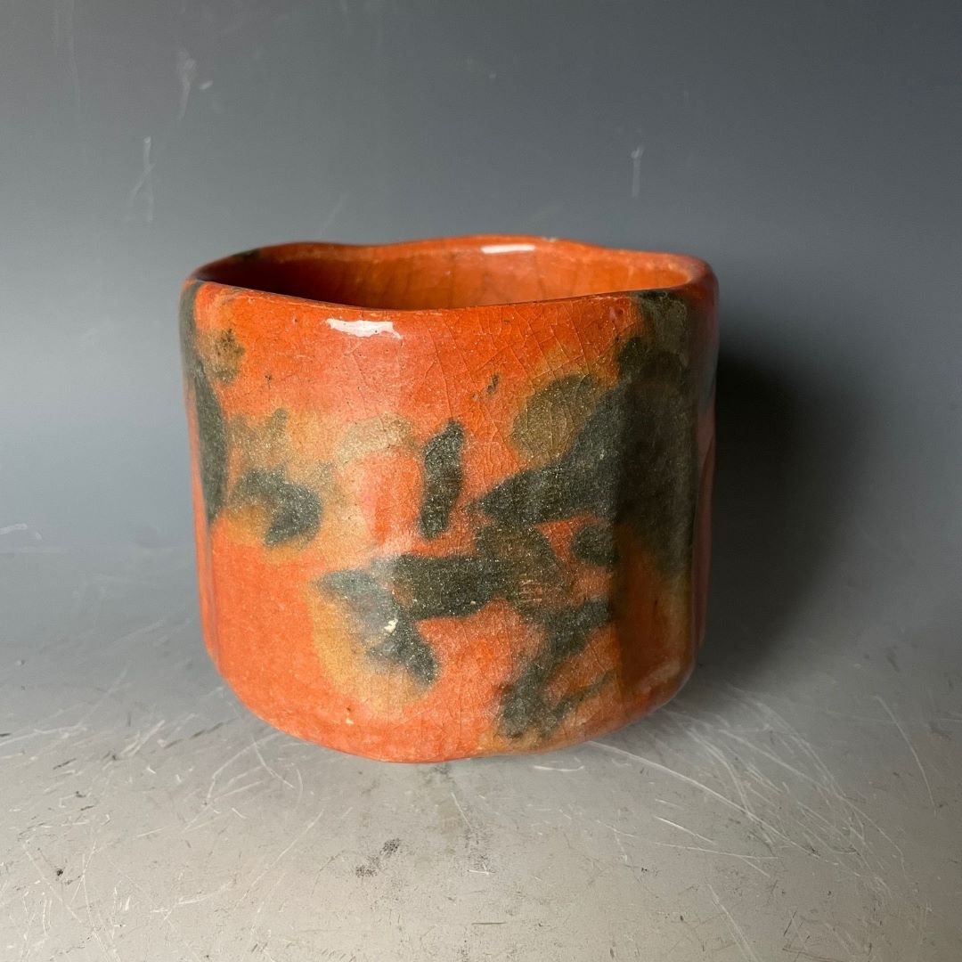 A traditional Japanese Akaraku-style Raku yaki tea bowl used in tea ceremonies, featuring a vibrant orange glaze with abstract black patterns, showcasing the unique pottery craftsmanship.