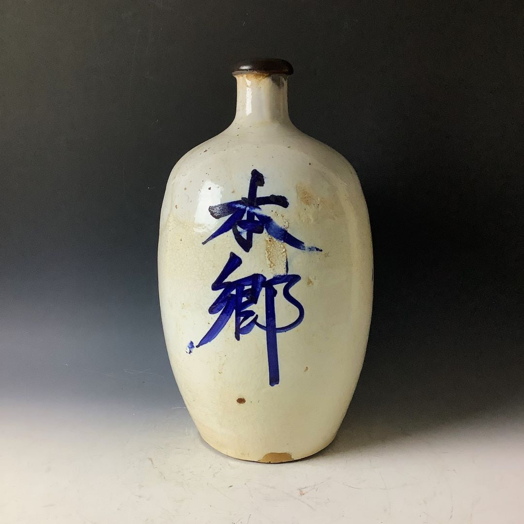 A Japanese Tokkuri sake bottle with vibrant blue kanji script on an off-white glaze, topped with a dark brown lip, set against a soft-lit black background.