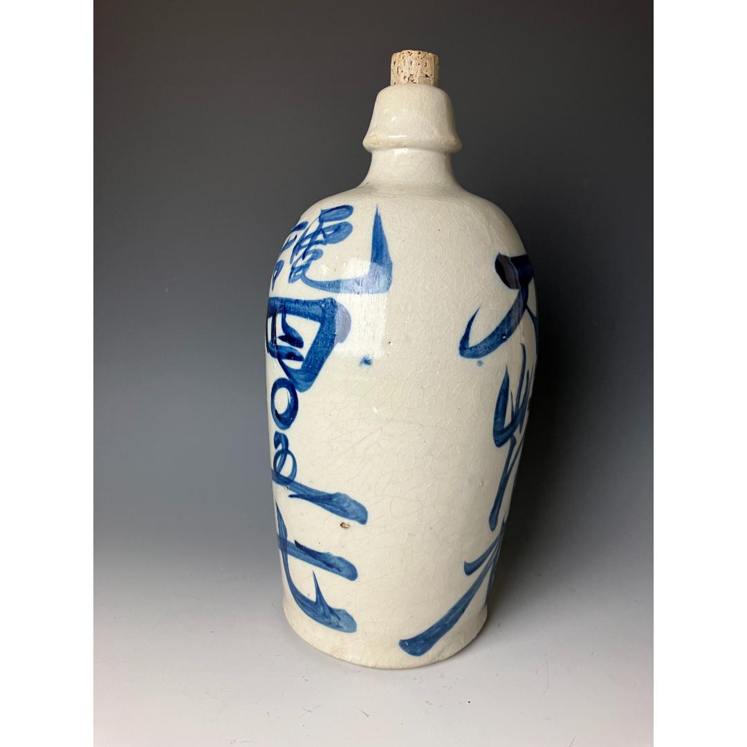 A side view of a Japanese ceramic sake bottle with bold blue kanji characters, featuring a textured white glaze and a cork stopper, set against a dark grey background.